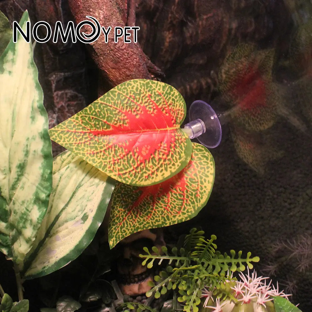 An artificial aquarium plant with a red and green patterned leaf attached to a clear suction cup, shown in a terrarium setting with other plants and rocks. The brand name 'NOMOYET' is visible in the top left corner.