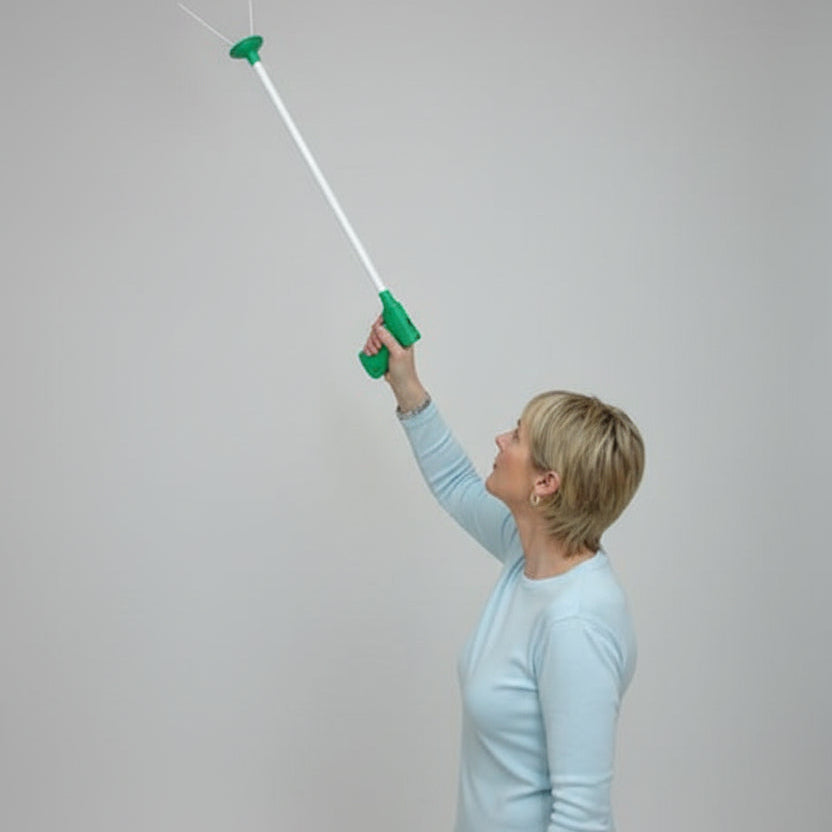 A woman wearing a white long-sleeve shirt and green gloves uses a long-handled paint roller to apply white paint to a room corner with beige walls and a wooden ceiling beam.
