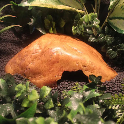 A natural-looking tan rock hide with a hollow center, displayed on dark gravel surrounded by green artificial plants.